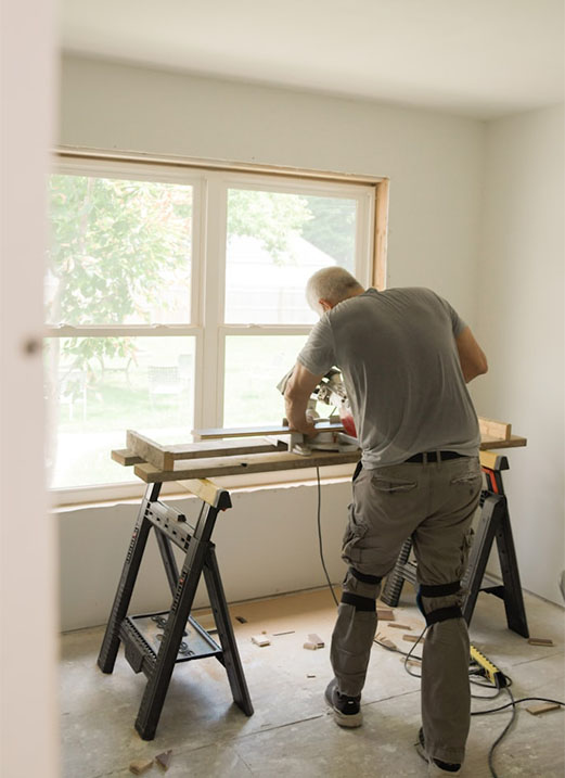 A man working at a wooden table saw in a bright, unfinished room with large windows, surrounded by sawdust.