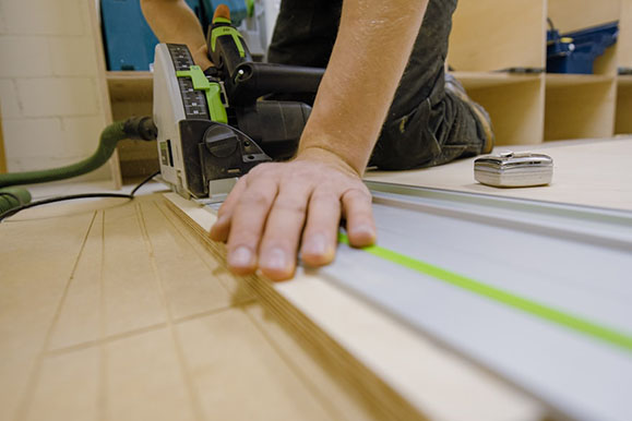 A close-up of a person's hand guiding a circular saw along a guide rail, focused on cutting plywood in a workshop setting.